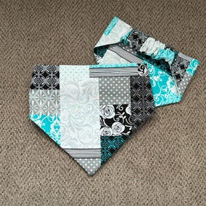 May include: A triangular pet bandana featuring a patchwork design of black, white, gray, and turquoise fabric squares. The patterns include floral swirls, polka dots, geometric shapes, and stripes. It has an elasticized neck band.