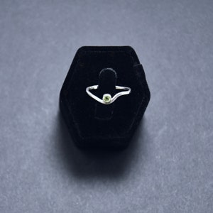 Puede incluir: Un anillo de plata con un diseño de onda y una pequeña piedra preciosa redonda de color verde claro. El anillo se exhibe sobre un soporte de anillo de terciopelo negro sobre un fondo gris oscuro.