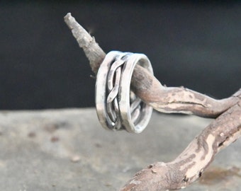 Könnte beinhalten: Ein silberner Ring mit einem geflochtenen Design. Der Ring wird auf einem kleinen, braunen Ast vor einem verschwommenen Hintergrund präsentiert. Die Textur und das komplizierte Muster des Rings werden in dieser Nahaufnahme hervorgehoben.