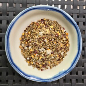 May include: A small white bowl with a blue rim contains a mixture of dried herbs, spices, and salt. The mixture is a blend of green herbs, red pepper flakes, and white salt crystals.