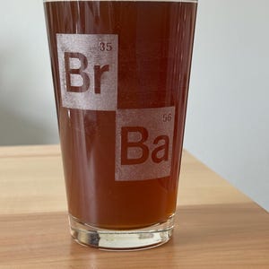May include: A clear glass pint with a brown liquid inside. The glass has the periodic table symbols "Br" and "Ba" etched on the front. The glass is sitting on a light-colored wooden surface.