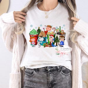 May include: White long-sleeved shirt with a Christmas-themed graphic featuring the Peanuts characters, including Charlie Brown, Snoopy and Lucy, holding various holiday-themed drinks. The graphic also includes snowflakes and a Christmas tree.