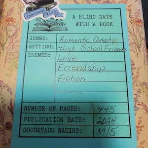 May include: A wrapped book with a floral pattern and a sticker reading I've lived a thousand lives. A light blue label reads A Blind Date With A Book, listing genre as Romantic Comedy, setting as High School Friends, themes as Love, Friendship, Fiction, 445 pages, 2024 date, and 3.9/5 rating.