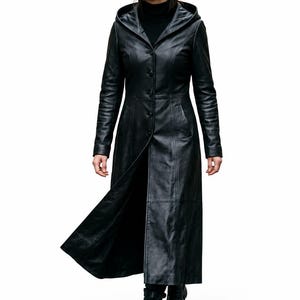 May include: A long black leather coat featuring a hood, a button-front closure, and long sleeves. The coat has a fitted bodice that flares slightly toward the hem, creating a structured silhouette. It is worn over a black turtleneck and black boots.