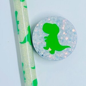 May include: A white pen with green dinosaur silhouettes and a white phone grip with a green dinosaur silhouette. Both items have a sparkly finish.