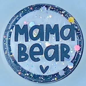 May include: A white round sticker with a black border and glitter embedded in the white surface. The sticker says "mama bear" in black block letters with a small heart below the text.