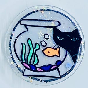 May include: A black cat peeks into a round fishbowl with a single orange fish, green seaweed, and blue and pink rocks. The fishbowl is decorated with white glitter.