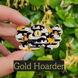 May include: A gold enamel pin depicting a stylized creature with a large yellow eye, black and white stripes, and cloud-like details. The pin has a gold edge and the text "Gold Hoarder" in a black box.