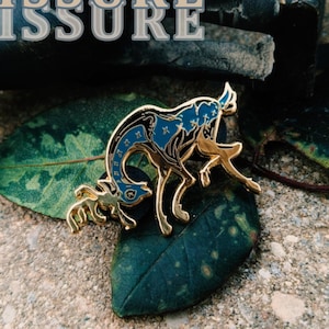 Może przedstawiać: Złota przypinka przedstawiająca dwa jelenie, jeden większy i jeden mniejszy, z niebiesko-złotym niebiańskim wzorem. Słowo "FISSURE" jest wyświetlane w wielu warstwach tekstu. Przypinka spoczywa na zielonym liściu i teksturowanej powierzchni.