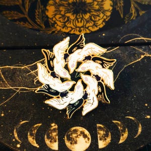 Peut inclure: Épingle circulaire en émail représentant des oiseaux blancs stylisés avec des ailes noires et des contours dorés. Les oiseaux sont disposés en cercle. L'épingle est posée sur un fond noir avec des motifs floraux et célestes dorés.