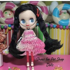 Puede incluir: Una muñeca con cabello negro en coletas, vestida con un vestido de ganchillo rosa y blanco. La muñeca tiene ojos azules y labios rojos. El texto "Shoes for Pet Shop Dolls" es visible en la parte inferior de la imagen.