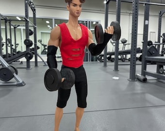 May include: A doll in a gym setting, lifting dumbbells. The doll is wearing a red sleeveless shirt with cutouts, black shorts, and black arm sleeves. The background shows gym equipment.