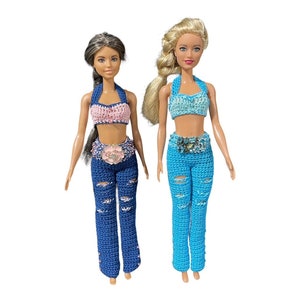 May include: Two dolls wearing crocheted outfits. One doll has blonde hair and is wearing a light blue crocheted top and trousers. The other doll has brown hair and is wearing a pink crocheted top and blue crocheted trousers.