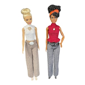 May include: Two dolls wearing crocheted outfits. One doll has blonde hair and is wearing a white crocheted top, a brown crocheted belt, and brown crocheted pants. The other doll has brown hair and is wearing a red crocheted top, a black crocheted belt, and gray crocheted pants.