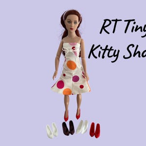 May include: A doll wearing a white dress with orange and purple polka dots, and red shoes. Several pairs of miniature shoes in white, brown, and red are at the doll's feet. The text "RT Tiny Kitty Shoes" is in the upper right corner.