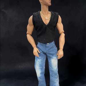 May include: A posable doll with brown hair styled in an undercut, wearing a black sleeveless V-neck top, a gold-toned chain necklace, and blue denim jeans with light-colored distressed patches on the thighs and knees, standing against a black background.