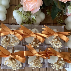 May include: Six small white cloud-shaped candles are displayed in clear boxes with gold ribbon and dried white flowers. The boxes are arranged in two rows of three.