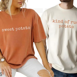 May include: Two t-shirts, one burnt orange and one cream, with the words "sweet potato" and "kind of rude potato" printed on them. The shirts are short-sleeved and appear to be made of a soft material. The shirts are part of a matching set.