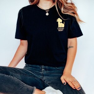 Może przedstawiać: Czarny t-shirt z żółtym kreskówkowym psem na lewej kieszeni na piersi. Tekst "Cute But Derpy" znajduje się pod psem.