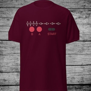 May include: A maroon t-shirt with a white graphic of a video game controller. The graphic includes the directional buttons, A, B, and Start buttons.