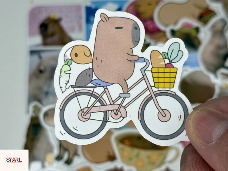 May include: A sticker featuring a cartoon capybara riding a pink bicycle with a yellow basket filled with bread and vegetables. A small companion sits on the back of the bike. The sticker has a white border.