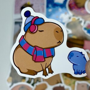 May include: A sticker featuring a cartoon capybara wearing a blue and pink striped scarf, hat, and earmuffs. A smaller blue capybara is next to the larger one. The sticker has a white background.