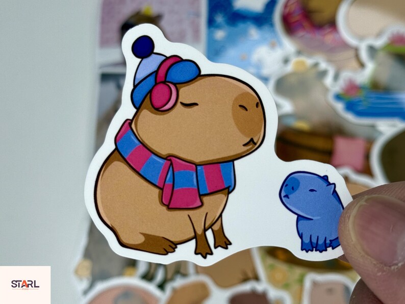 May include: A sticker featuring a cartoon capybara wearing a blue and pink striped scarf, hat, and earmuffs. A smaller blue capybara is next to the larger one. The sticker has a white background.
