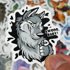 May include: A sticker featuring a cartoon wolf with gray and white fur, a smug expression, and the text "AWOOO, BABY!". The wolf is set against a black and white splash design.