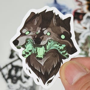 May include: A sticker featuring a three-headed wolf illustration. The wolves are depicted in shades of brown and gray, with glowing green eyes and a green, monstrous maw. The sticker has a white border and a stylized design.