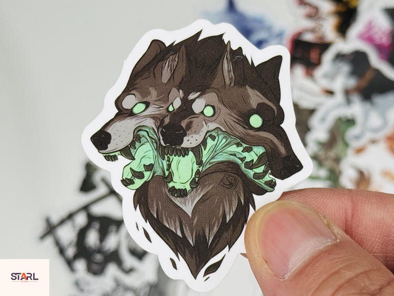 May include: A sticker featuring a three-headed wolf illustration. The wolves are depicted in shades of brown and gray, with glowing green eyes and a green, monstrous maw. The sticker has a white border and a stylized design.