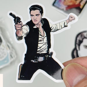 May include: A sticker featuring a black and white illustration of a man with dark hair wearing a black vest and a white shirt. He is holding a gun in his right hand and has a serious expression on his face.