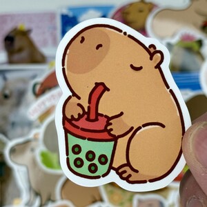 May include: A sticker featuring a cartoon capybara drinking from a boba tea cup. The capybara is light brown with a darker brown head and is holding a red and green cup with green boba. Other capybara stickers are in the background.
