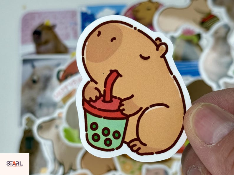 May include: A sticker featuring a cartoon capybara drinking from a boba tea cup. The capybara is light brown with a darker brown head and is holding a red and green cup with green boba. Other capybara stickers are in the background.