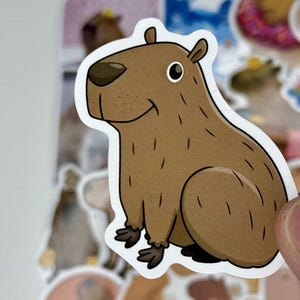 May include: A sticker featuring a cartoon capybara in shades of brown, with a white outline. The capybara is depicted in a sitting position, with a friendly expression. The sticker is held by a person's fingers, with other stickers in the background.