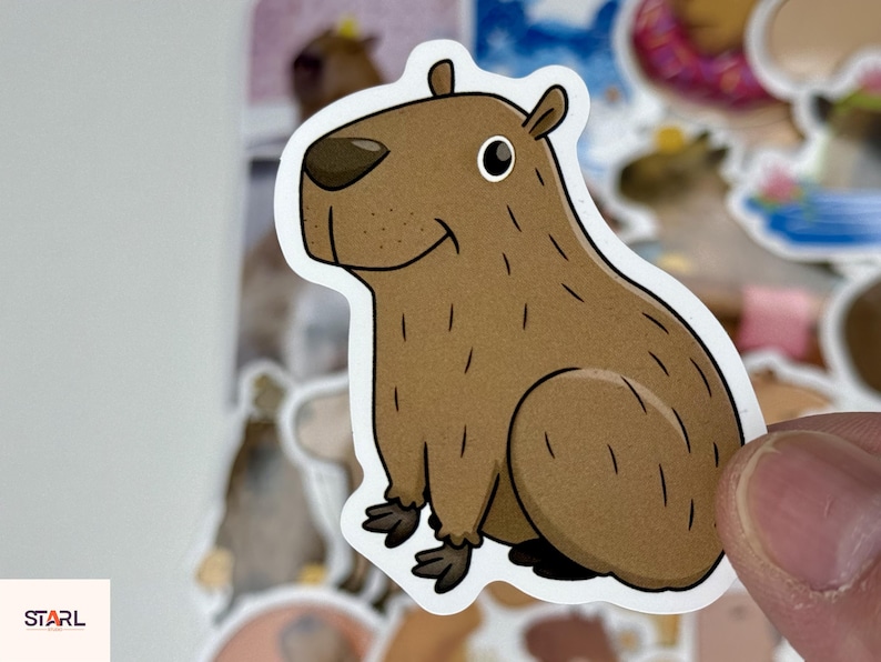 May include: A sticker featuring a cartoon capybara in shades of brown, with a white outline. The capybara is depicted in a sitting position, with a friendly expression. The sticker is held by a person's fingers, with other stickers in the background.