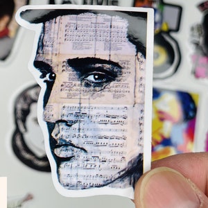 May include: Close-up of a sticker featuring a black and white portrait of a man with a sheet of music behind him. The man's eye is visible and the sheet music is faded and has a vintage look.
