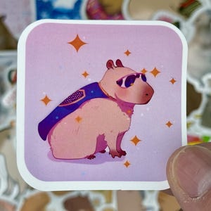 May include: A square sticker featuring a cartoon capybara wearing sunglasses and a superhero cape. The capybara is light brown with a purple cape and sunglasses. Orange starbursts are scattered around the capybara on a pink background. The sticker is held by a hand.