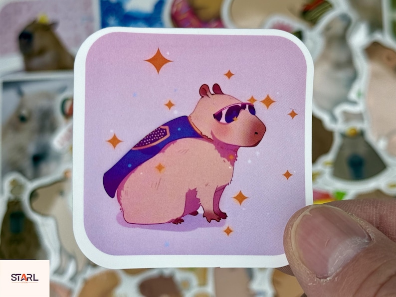 May include: A square sticker featuring a cartoon capybara wearing sunglasses and a superhero cape. The capybara is light brown with a purple cape and sunglasses. Orange starbursts are scattered around the capybara on a pink background. The sticker is held by a hand.
