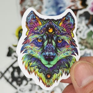 May include: A colorful wolf sticker with intricate details. The wolf's face is depicted with vibrant hues of blue, purple, green, and yellow. The sticker has a white border and is held by a hand. The word "STARL" is visible in the lower left corner.