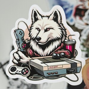 May include: A sticker featuring a white wolf with pink eyes holding a retro video game controller. The wolf is next to a console, a drink, and another controller. The sticker has a retro gaming theme.