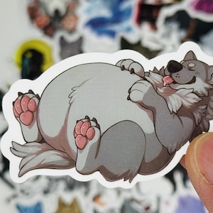 May include: A sticker featuring a cartoon wolf lying on its back with its tongue out. The wolf has gray fur, pink paws, and a happy expression. The sticker is white with a rounded edge. Other stickers are visible in the background.