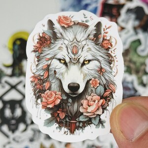 May include: A sticker featuring a detailed illustration of a wolf's face, adorned with roses and foliage. The wolf has yellow eyes and a decorative headpiece. The sticker has a white border and is held by a hand.