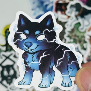 May include: A sticker featuring a cartoon dog with a black and blue color scheme, highlighted by white lightning bolt accents. The dog has glowing white eyes and a friendly expression. The sticker is held by a hand, with other stickers in the background.