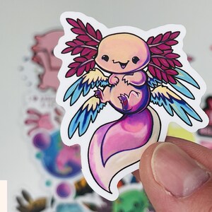 Op de afbeelding: Een sticker met een cartoon-axolotl met roze, blauwe en gele vleugels. De axolotl is roze en wit met een glimlachend gezicht.