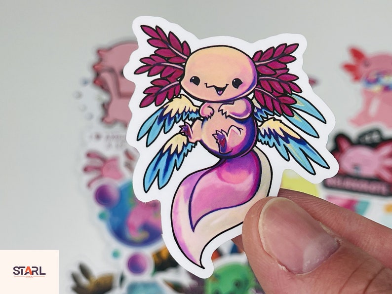 Op de afbeelding: Een sticker met een cartoon-axolotl met roze, blauwe en gele vleugels. De axolotl is roze en wit met een glimlachend gezicht.