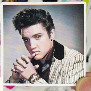 May include: A black and white photo of Elvis Presley wearing a striped jacket and a dark shirt. He is looking at the camera and has his hand on his chin.