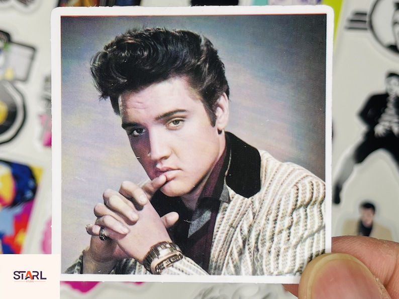 May include: A black and white photo of Elvis Presley wearing a striped jacket and a dark shirt. He is looking at the camera and has his hand on his chin.
