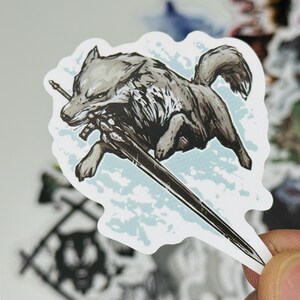 May include: A sticker featuring a gray wolf with a sword in its mouth, running across a light blue background. The wolf is detailed with black and white shading. The sticker has a white border and is surrounded by other stickers.