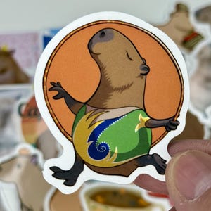 May include: A sticker featuring a cartoon capybara with a brown body, black feet and arms, and a green and yellow patterned shirt. The capybara is set against an orange circle.
