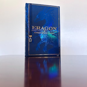 May include: A hardcover book titled Eragon by Christopher Paolini with a deep blue cover featuring a large, embossed dragon illustration in metallic blue and gold foil accents, complete with a dark blue ribbon bookmark and a small oval charm.